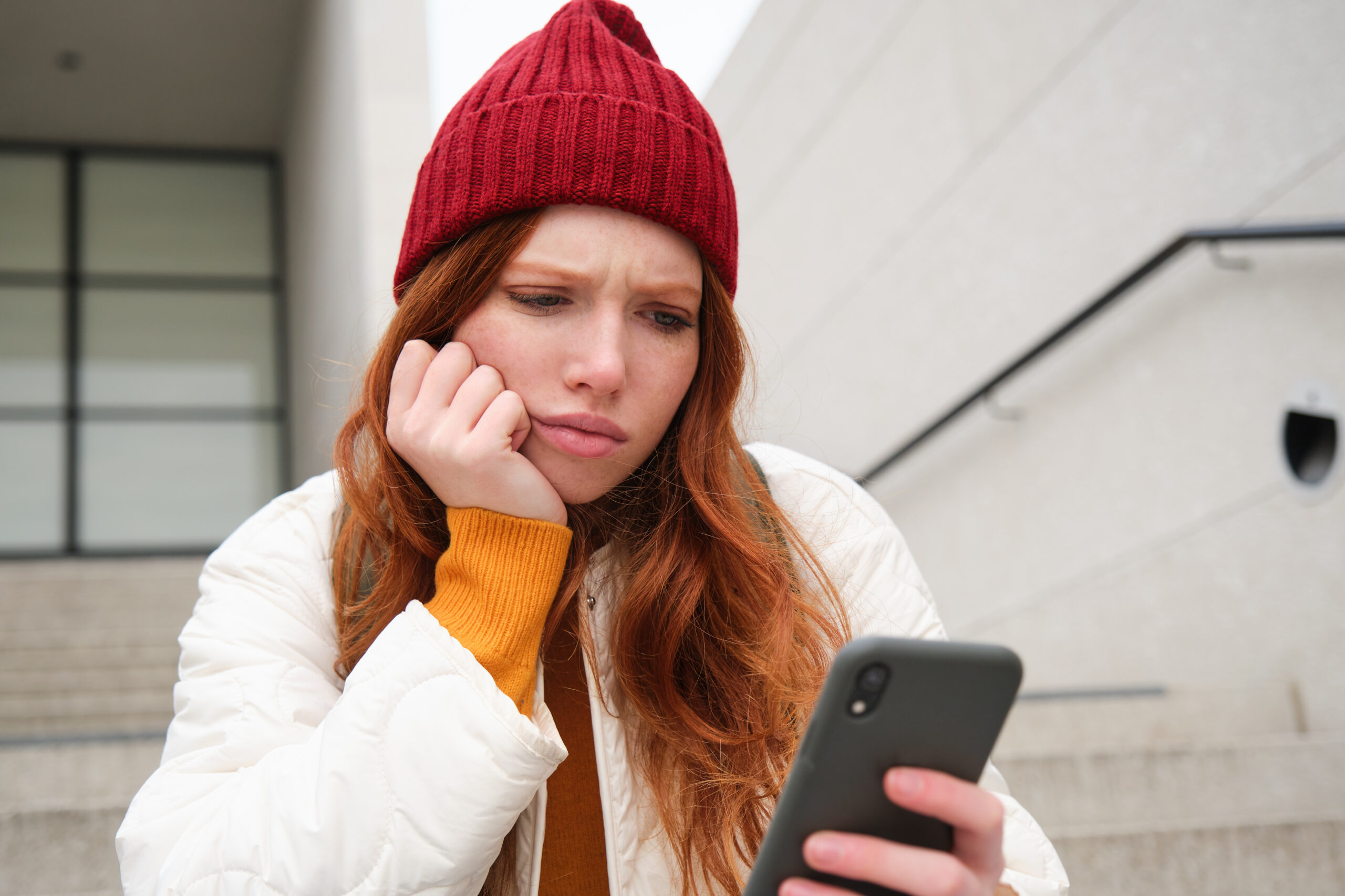 Portrait of sad redhead girl, looks upset and disappointed at smartphone screen, reads bad news text message on mobile phone and frowns, sits on stairs outdoors.